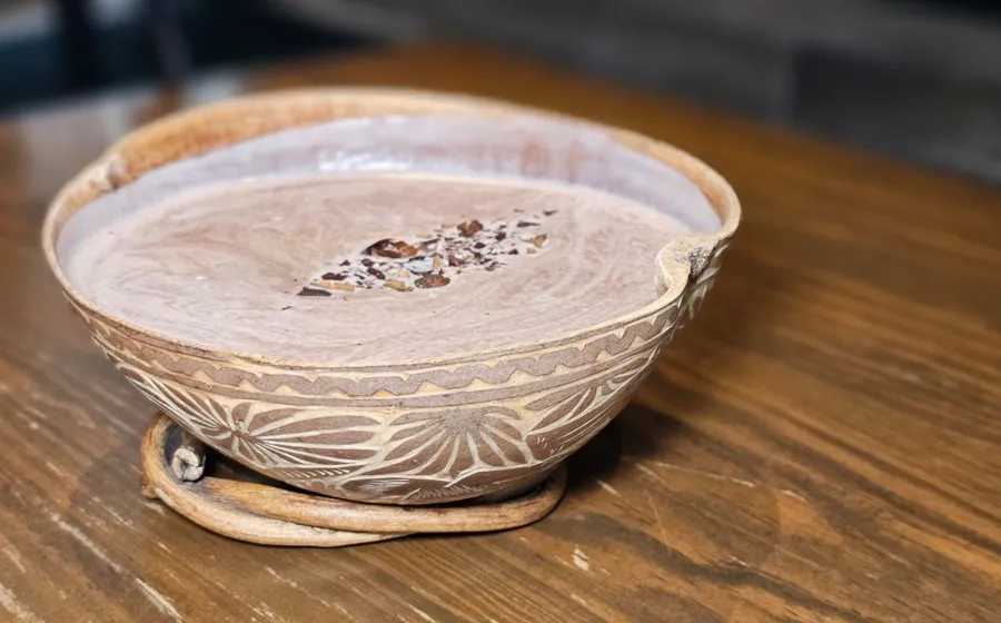 Bebida de cacao con nibs servida en una jícara tallada sobre una mesa de madera.